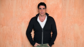 Hombre de cabello oscuro con chaqueta negra de cremallera y camiseta blanca ante una pared anaranjada.