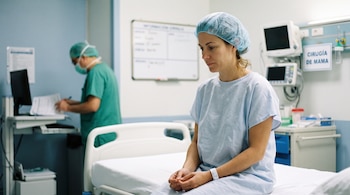 Una mujer con bata y gorro quirúrgico sentada en una cama de hospital, junto a un sanitario con uniforme verde y monitores médicos.