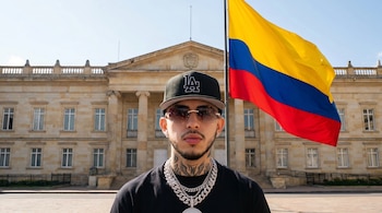 Hombre joven con gorra, gafas de sol y collares de plata posa frente a un edificio colonial y una bandera de Colombia a la derecha, bajo un cielo claro.