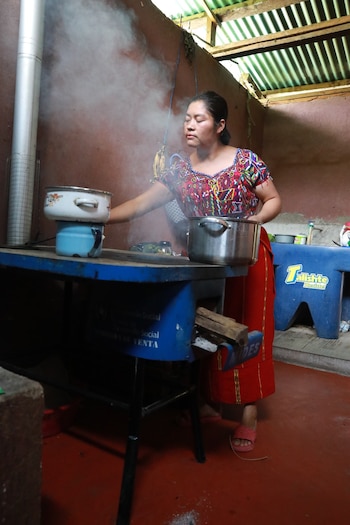 Mujer con blusa colorida y falda roja cocina en un fogón rústico con humo, manejando ollas de metal y barro sobre una mesa azul