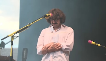 Un hombre con cabello largo, gafas y bigote, vestido con camisa clara, tiene las manos juntas en un escenario con dos micrófonos amarillos y rosados