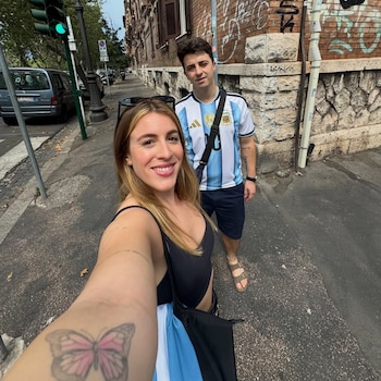 Mujer rubia sonriente en primer plano con tatuaje de mariposa. Hombre joven detrás con camiseta de fútbol de Argentina. Calle con edificios y semáforo