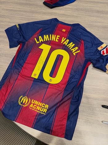 Una camiseta de fútbol del FC Barcelona con franjas azules y rojas, el nombre Lamine Yamal, el número diez autografiado, y el logo de ACNUR