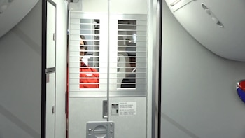Una puerta metálica con rejillas instalada en el pasillo de un avión, con una auxiliar de vuelo y un piloto detrás de la estructura