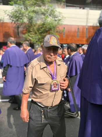 Un hombre de edad avanzada con gorra y un escapulario al cuello camina junto a fieles vestidos con hábitos morados en una calle