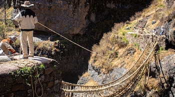 Un hombre con sombrero sostiene una soga sobre una estructura de piedra junto a un puente de fibra vegetal construido sobre un cañón.
