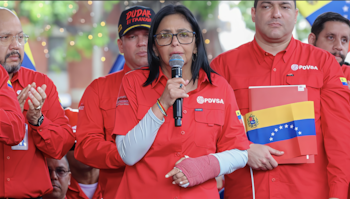 Una mujer con gafas, cabello oscuro y un yeso en el brazo izquierdo habla por un micrófono, vestida con una camisa roja, rodeada de hombres