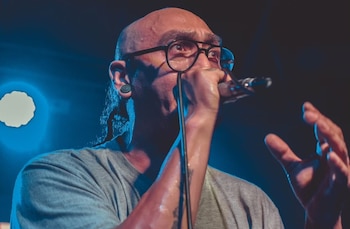 Un hombre calvo con gafas y camiseta gris canta en un micrófono. Tiene perforaciones en las orejas y gesticula con la mano derecha. Iluminación azul