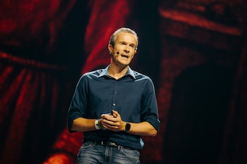 Hombre de mediana edad con cabello canoso y camisa azul oscuro habla en un escenario oscuro con luces rojas de fondo. Lleva micrófono de diadema y pulseras