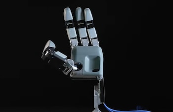 Una mano robótica de color gris y metálico con tres dedos extendidos y un pulgar sobre un fondo negro con cables azules en la base