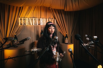 Una mujer con cabello oscuro y flequillo, vestida con chaqueta peluda y diadema rosa, parada frente a múltiples micrófonos y botellas de Fernet Branca