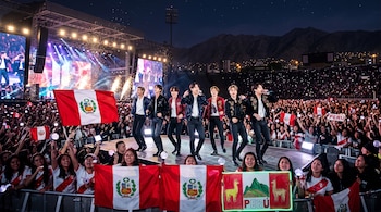 Los integrantes de BTS cantan en un escenario frente a una multitud que ondea banderas peruanas en un estadio al aire libre.