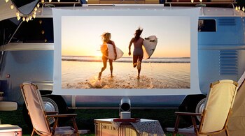 Furgoneta azul, pantalla blanca, proyector, mesa, dos sillas, luces decorativas, dos mujeres con tablas de surf en una playa al atardecer