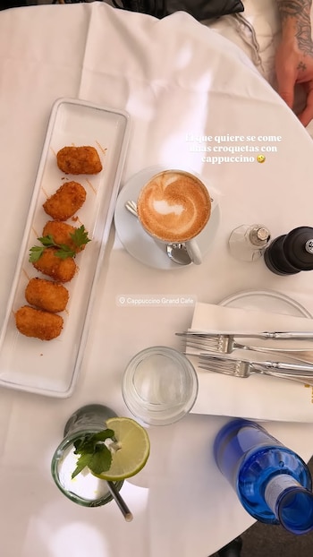 Vista aérea de una mesa de restaurante con un plato de croquetas, una taza de capuchino, un vaso con una bebida de lima y menta, y una botella azul