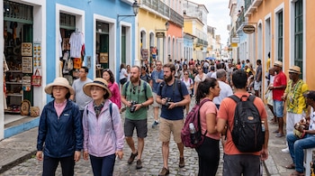 Una calle adoquinada con edificios coloniales de colores, personas caminando, algunas con cámaras, y tiendas abiertas en un día soleado.