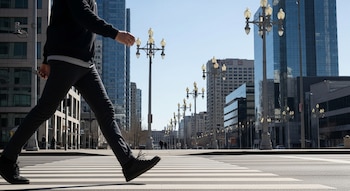 Primer plano de una persona caminando sobre un paso peatonal; al fondo, edificios de cristal y metal, y una hilera de farolas.