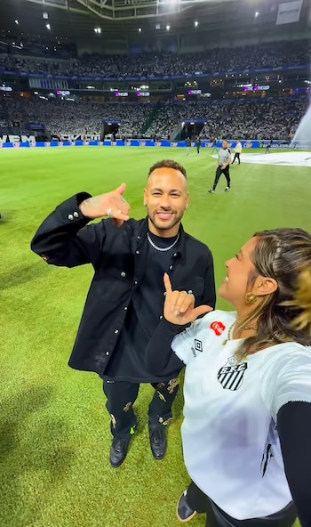 Neymar viste ropa negra y hace un gesto con la mano junto a una mujer con la camiseta blanca del Santos FC en un campo de fútbol