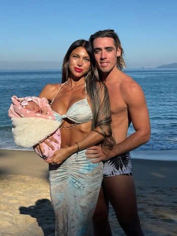 Cielo López sostiene a un bebé envuelto en una manta rosa junto a Claudio Spinelli en una playa frente al mar