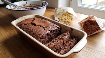 Budín de chocolate horneado en molde, rebanado. Un bol con masa, un batidor y dos bolsas con quinoa y cacao en polvo sobre mesa de madera junto a ventana.