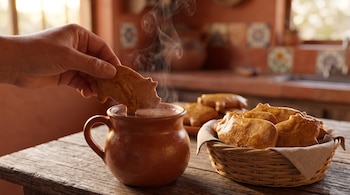 Una mano introduce una galleta en forma de cerdo en una taza de barro humeante, al lado de una cesta con galletas sobre una mesa de madera.