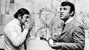Fotografía en blanco y negro de dos hombres. Uno con camisa y corbata come. El otro con traje sostiene un vaso y un pañuelo, con fondo de azulejos.