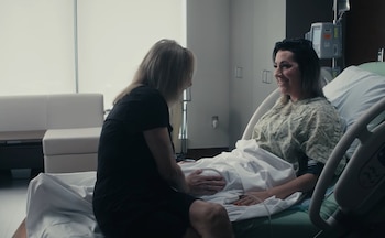 Dos mujeres en una habitación de hospital; una está recostada en la cama con bata y la otra permanece sentada a su lado