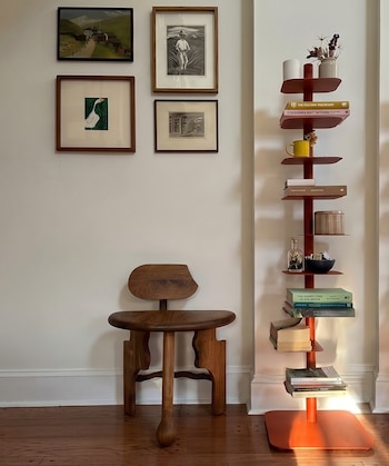 Vista de un espacio interior con paredes blancas, suelo de madera, una silla de madera, cuatro cuadros enmarcados y dos estanterías rojas con libros y objetos.