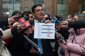 Hombre con gafas y bigote, de traje y corbata, sostiene un documento con tablas de datos frente a micrófonos de prensa. Al fondo, se ven más personas y un edificio