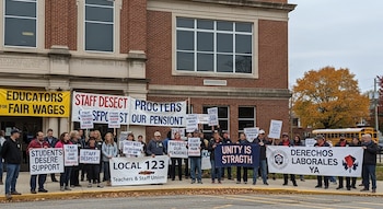 Grupo de docentes y personal escolar protestando con pancartas frente a un edificio de ladrillo rojo. Hay un autobús escolar amarillo y un árbol otoñal.