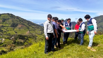Grupo de siete hombres, algunos con chalecos de seguridad, observando un plano en una ladera cubierta de hierba con montañas verdes al fondo bajo un cielo azul claro