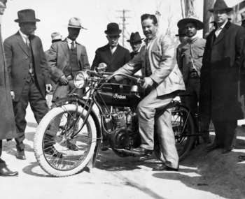 Francisco Villa, sentado en una motocicleta Indian de época con neumáticos blancos, rodeado de varios hombres vestidos con trajes y sombreros en un camino de tierra