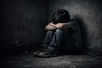 Niño sentado en el suelo de un rincón oscuro. Viste camiseta gris, pantalón vaquero y zapatillas oscuras. Su cabeza se apoya en los brazos cruzados.