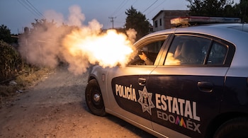 Patrulla de policía del Estado de México con destello y humo saliendo de la ventana del conductor. Brazo y mano sujetan un arma.