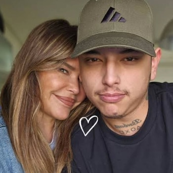 Mujer con cabello largo castaño claro, hombre joven con gorra verde, bigote y barba, y tatuaje en el cuello. Un corazón blanco se superpone a la imagen