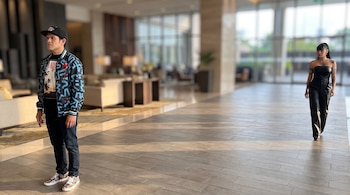 Un hombre con gorra, chaqueta estampada y jeans, y una mujer con top sin tirantes y pantalones, posan en un vestíbulo de hotel con suelo de baldosas.