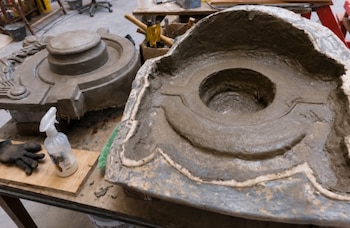 Dos piezas de molde de color gris, una llena de material húmedo y otra vacía, junto a un pulverizador blanco, un guante oscuro y herramientas sobre una mesa