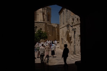 Un grupo de personas camina por una calle empedrada y soleada enmarcada por un pasaje oscuro, con edificios de piedra antiguos y una torre redonda visible