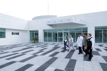 Vista exterior de un edificio blanco moderno con grandes ventanas de cristal. Cinco personas caminan sobre un pavimento de bloques grises y negros