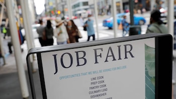 Un cartel negro con letras blancas que dicen "JOB FAIR" y una lista de puestos de cocina, en el primer plano, con personas borrosas caminando en una calle urbana detrás