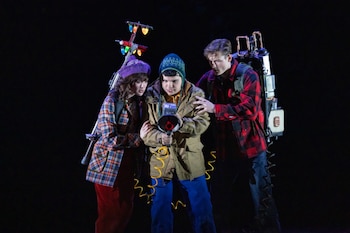 Tres jóvenes, dos hombres y una mujer, con gorros y chaquetas. Un hombre sostiene un megáfono. Una mochila con luces de Navidad y otra con estructura metálica. Fondo oscuro