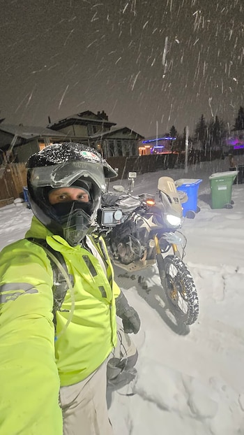 Un hombre con casco y chaqueta amarilla fluorescente está de pie junto a una motocicleta en una calle nevada. Nieve visiblemente cae de noche