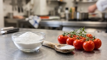 Recipiente de vidrio con polvo blanco, cuchara de madera y una rama de tomates rojos sobre una superficie metálica de cocina.