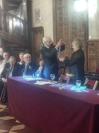 Angela Pradelli, con cabello rizado, recibe una condecoración de un hombre vestido de traje. Están frente a una mesa con mantel burdeos. Fondo de madera tallada