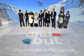 Diez personas, incluyendo personajes de orca y dinosaurio, un buzo y un conductor, posan en un escenario con fondo de montañas nevadas y el logo de Chubut