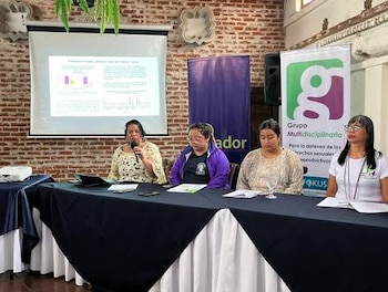 Cuatro mujeres sentadas tras una mesa con un micrófono frente a una pantalla de proyección y pancartas institucionales