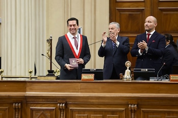 Hombre con traje y banda bicolor, sonriente, sostiene un libro. Dos hombres de traje a su lado. Podio de madera con campana, mazo y letrero de "1ra VICEPRESIDENCIA"