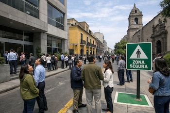 Una calle con personas evacuando edificios modernos y coloniales. Un letrero verde prominente indica "ZONA SEGURA" al centro. Cielo parcialmente nublado.