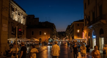 Una calle concurrida de noche en una ciudad europea con edificios iluminados, farolas, personas, un autobús, coches y un semáforo en rojo.