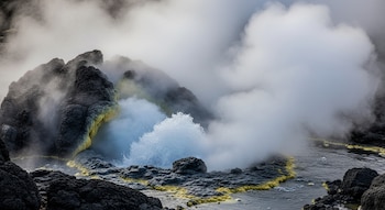 Detalle de agua burbujeante y vapor saliendo entre rocas volcánicas oscuras con minerales amarillos visibles.