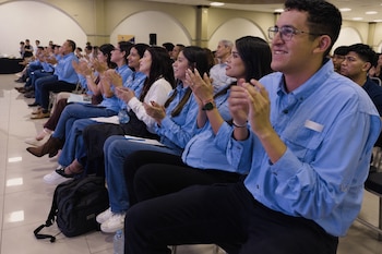 Fila de personas sentadas en un salón que aplauden vistiendo camisas de color azul claro y ropa casual.